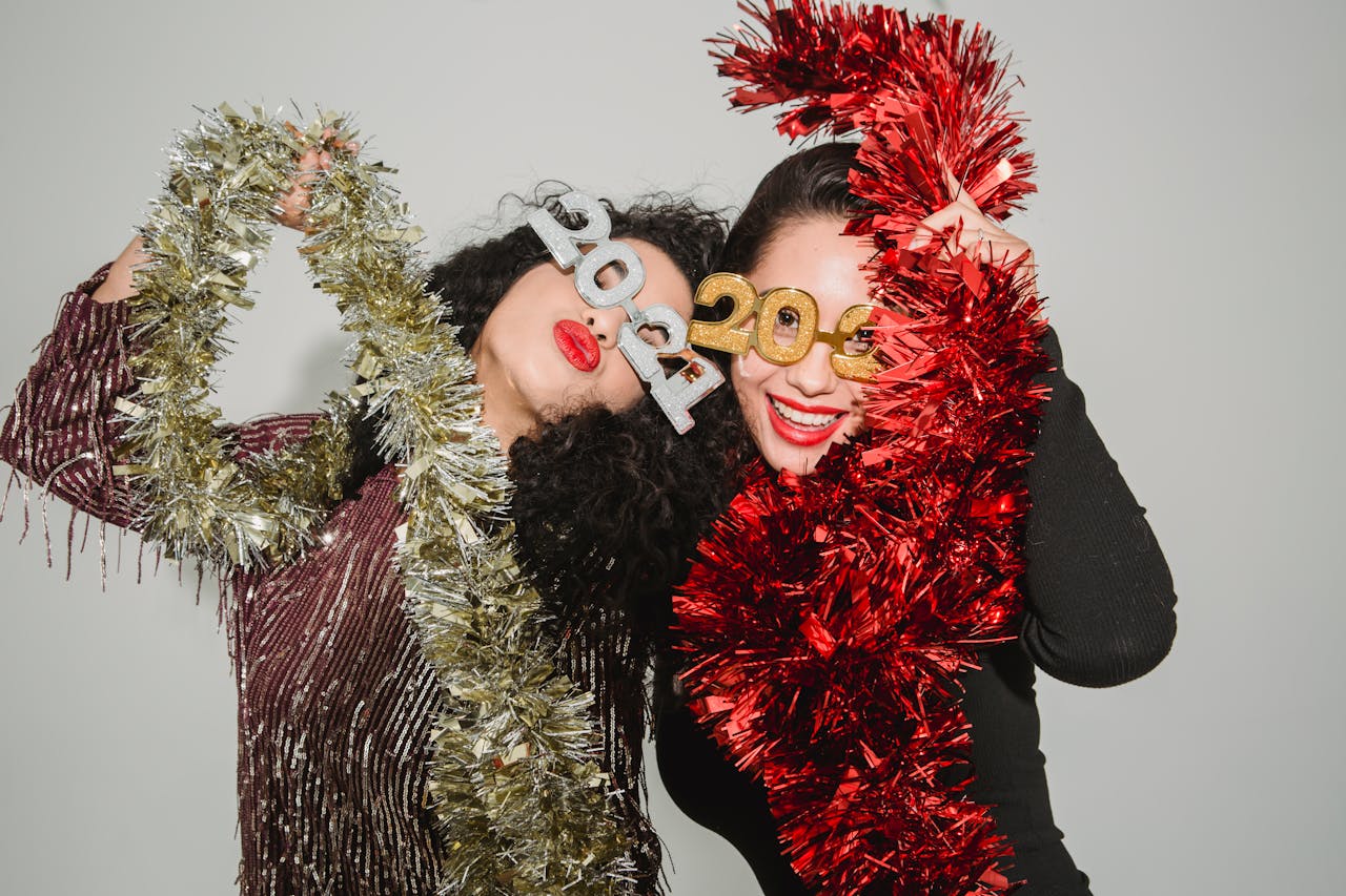 Happy women wearing festive dresses and 2021 glasses wrapped with shiny tinsel while celebrating New Year