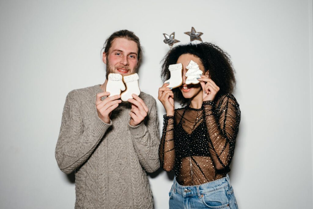 Smiling couple holding Christmas cookies, celebrating indoors with festive attire.